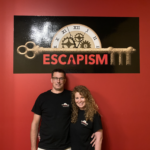 A man and a woman arm in arm, standing under the sign for the escape room.
