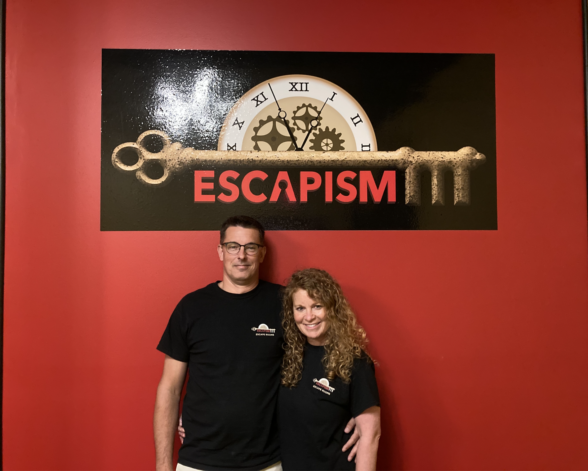 A man and a woman arm in arm, standing under the sign for the escape room.